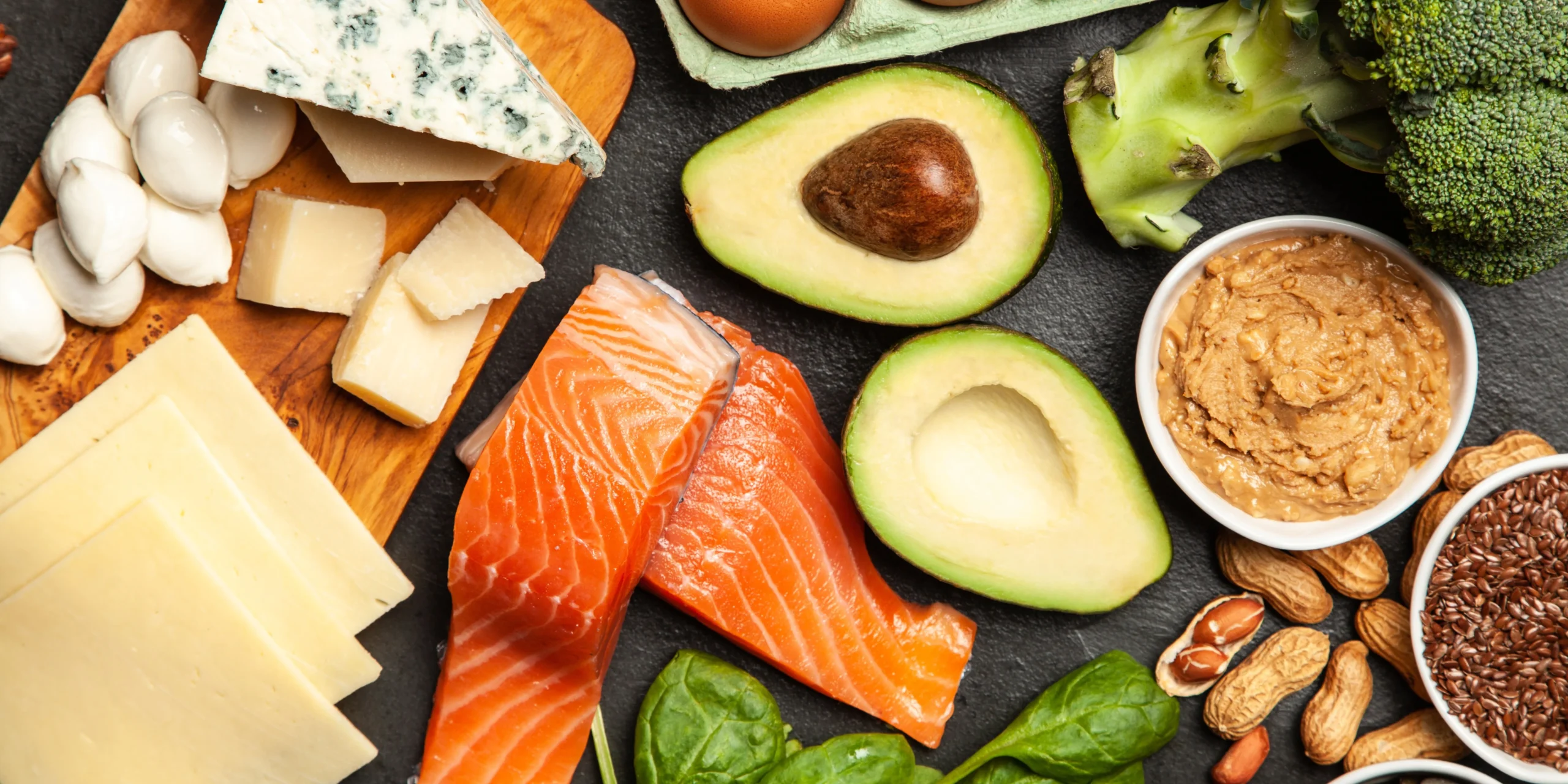 Alimentos saludables para dieta keto como salmón, aguacate, queso, brócoli y frutos secos sobre fondo oscuro.